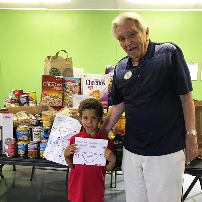 Ed Hughes posing with a child holding a thank you card in front of food at the Dunedin Cares Food Pantry